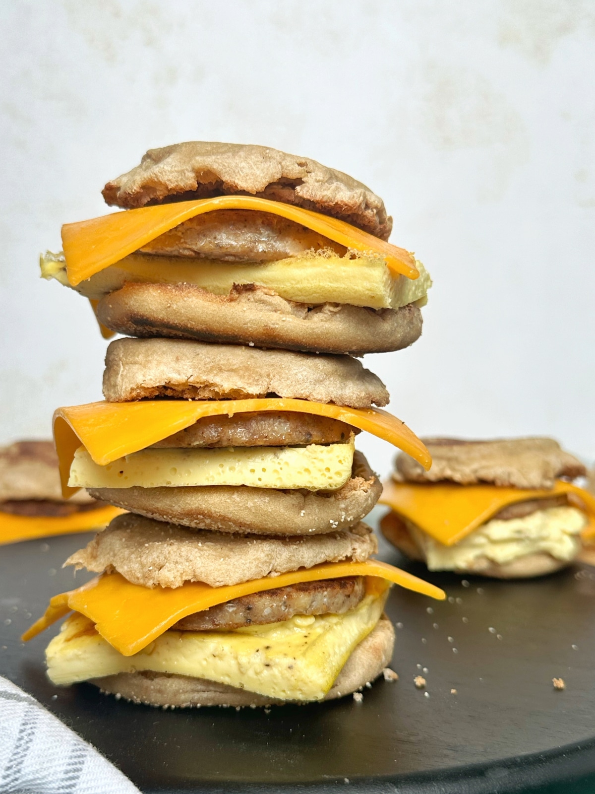 A stack of three sausage egg and cheese breakfast sandwiches on English muffins on a black wooden board.
