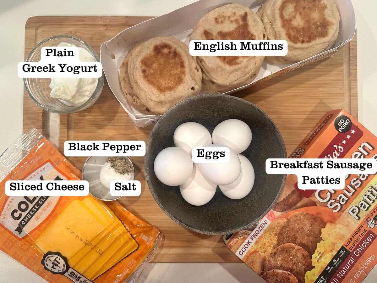 A wooden board containing ingredients for breakfast sandwiches, including English muffins, sliced cheese, eggs, and sausage patties.