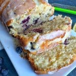 A loaf of lemon blueberry cake with lemon glaze, sliced on a white plate.