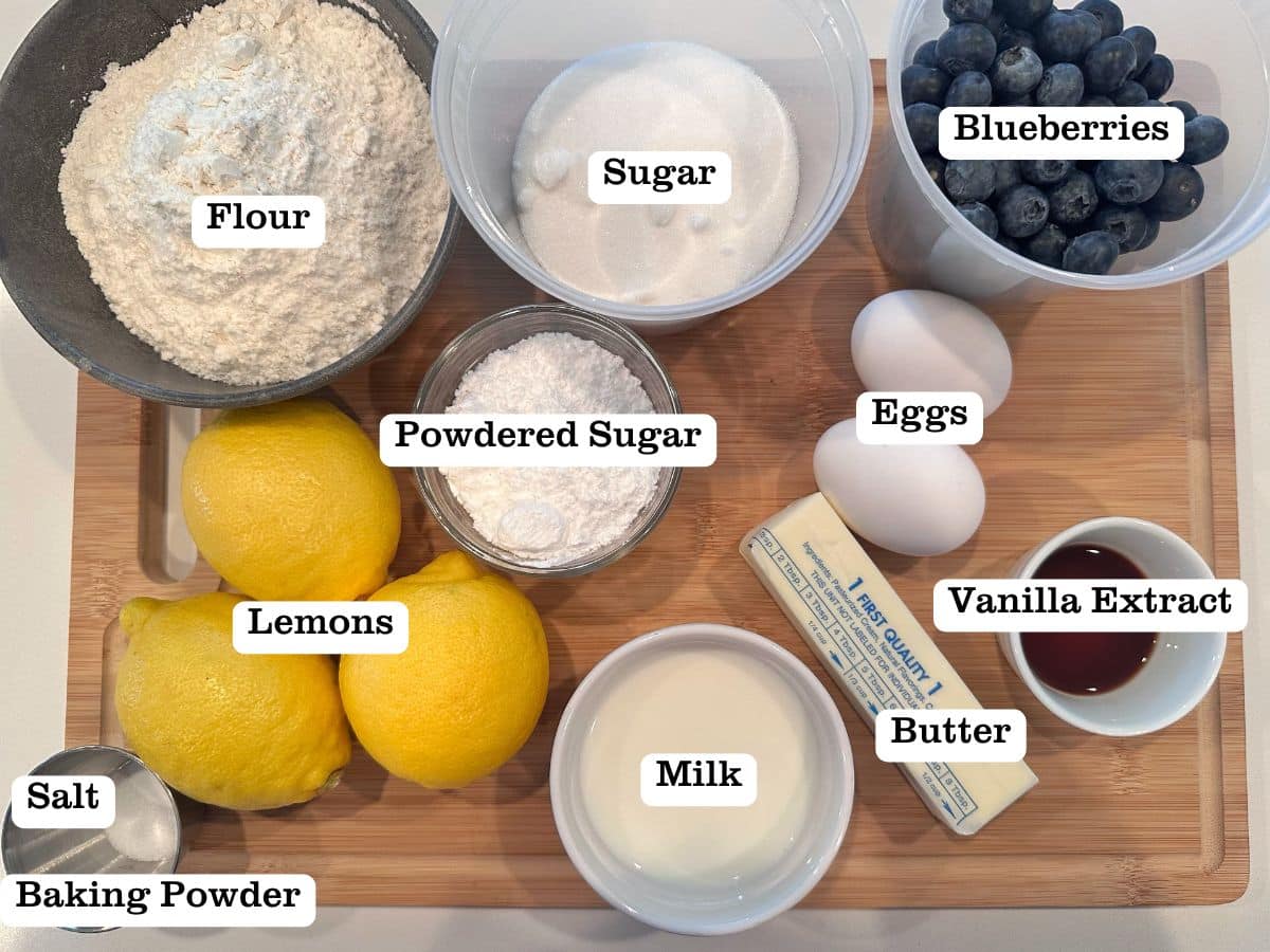 A wooden board containing ingredients for lemon blueberry cake, including flour, milk, powdered sugar, and fresh fruit.