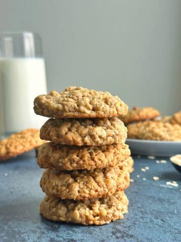 A stack of Chewy Oatmeal Cookies with a glass of milk and a plate full of extra cookies in the background.