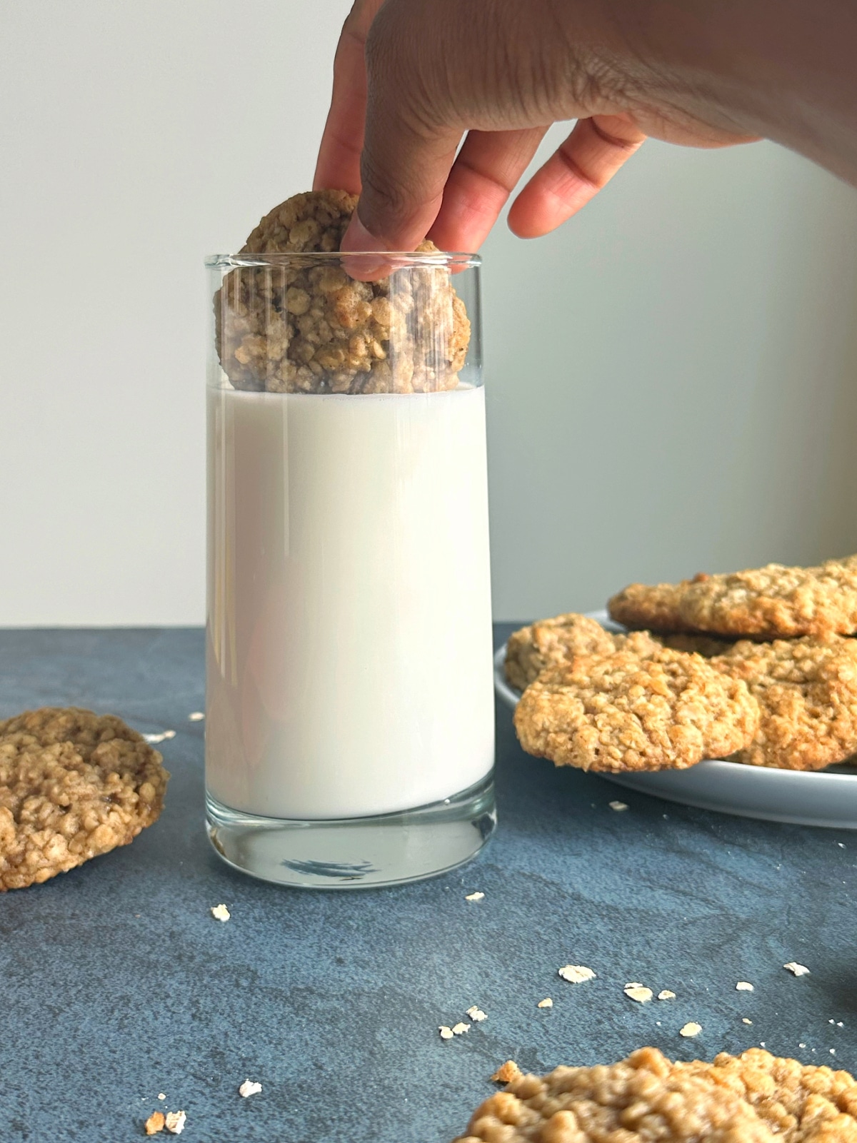 A hand dipping an oatmeal cookie into a tall glass of milk, with a plate of cookies in the background.