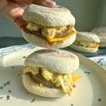 A hand grabbing the top sandwich of a stack of sausage, egg, and cheese breakfast sandwiches, with more in the background.