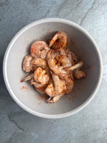 A bowl of shrimp marinating in seasonings like salt, pepper, and paprika.