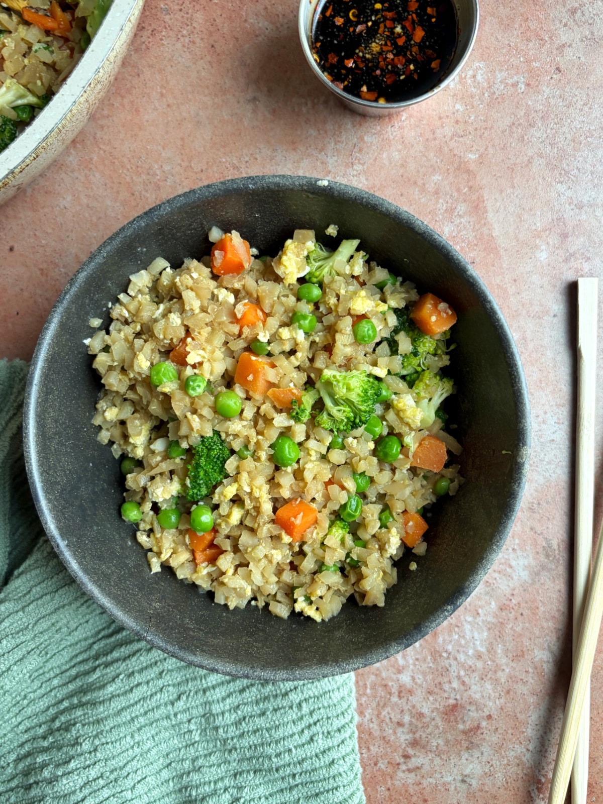 A black bowl filled with cauliflower fried rice, with chopsticks and extra soy sauce on the side.