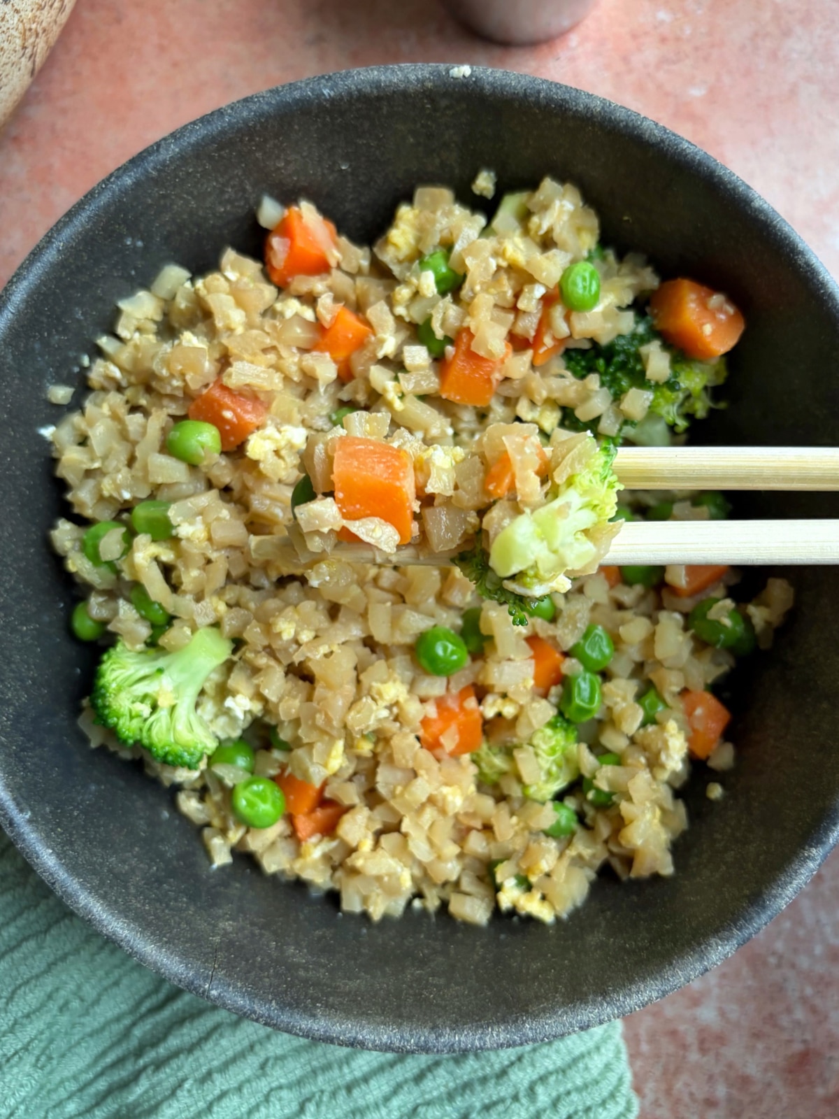 A pair of chopsticks holding up a bite of cauliflower fried rice over the bowl.