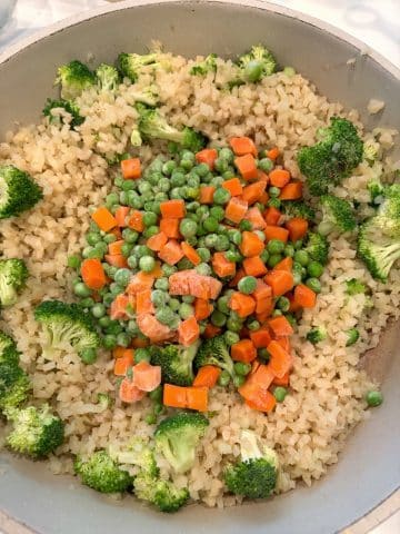 A skillet with riced cauliflower, frozen peas and carrots, and broccoli crowns inside.