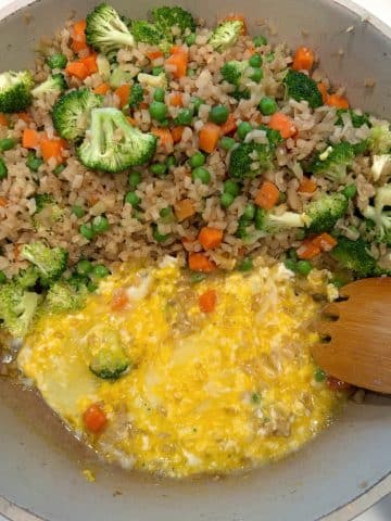 Eggs being scrambled by a wooden spatula in a skillet with cali-fried rice inside.