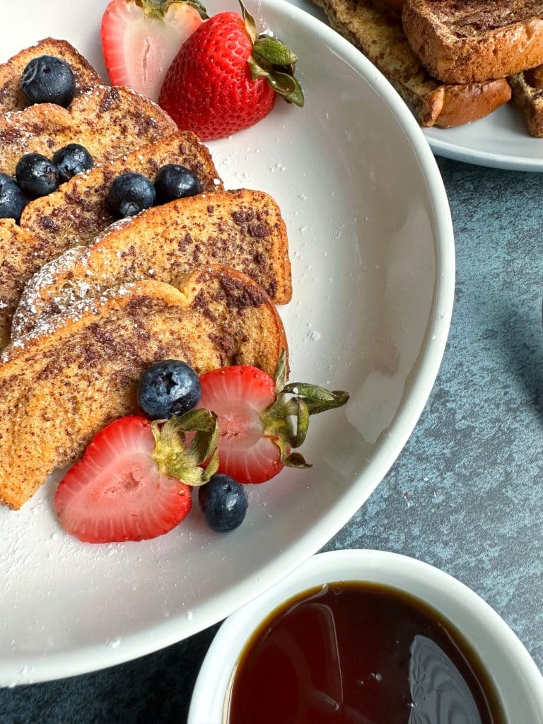 A closeup shot of a plate of baked French toast sticks with berries and powdered sugar.