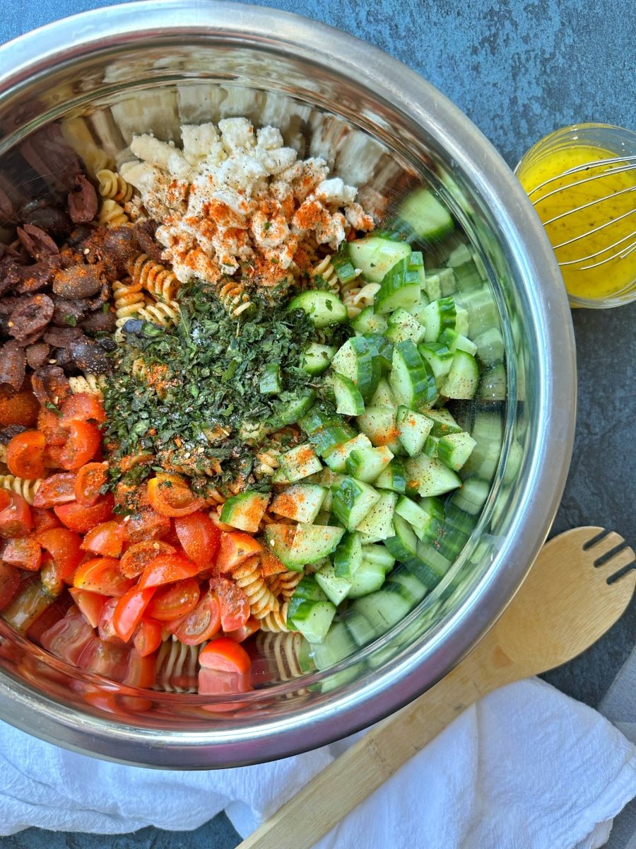 A bowl filled with the ingredients to mediterranean pasta salad - tomatoes, feta cheese, cucumbers, olives, and protein pasta.