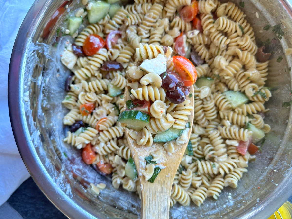 A wooden spoon containing a scoop of High Protein Mediterranean Pasta Salad with the rest of the bowl in the background.