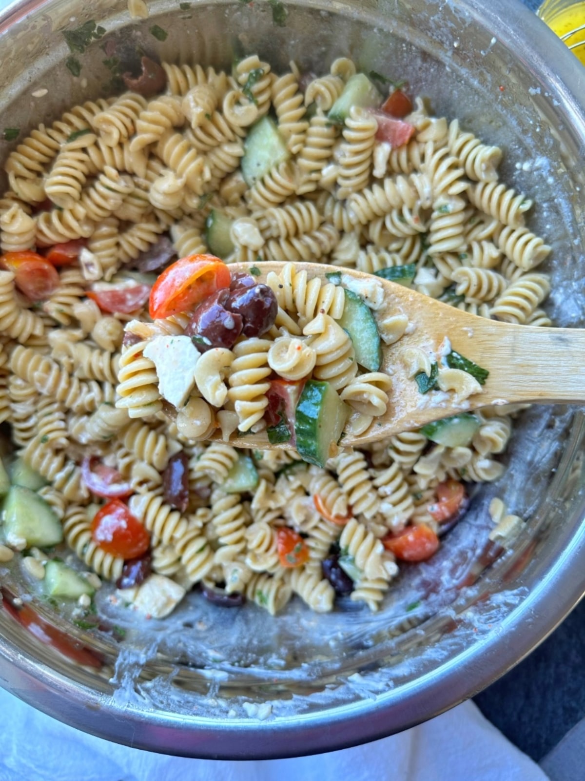 A spoon showing off a scoop of greek pasta salad over the remaining portions in a silver bowl.