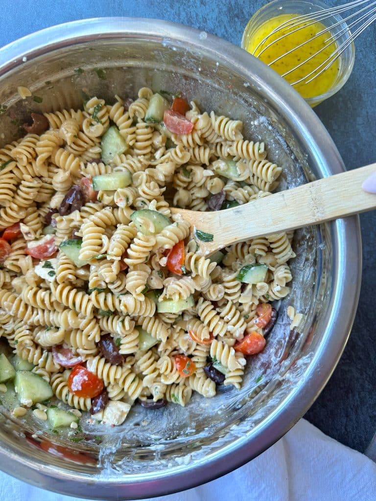 A large bowl of Mediterranean Pasta Salad mixed together with a wooden spoon in the bowl.