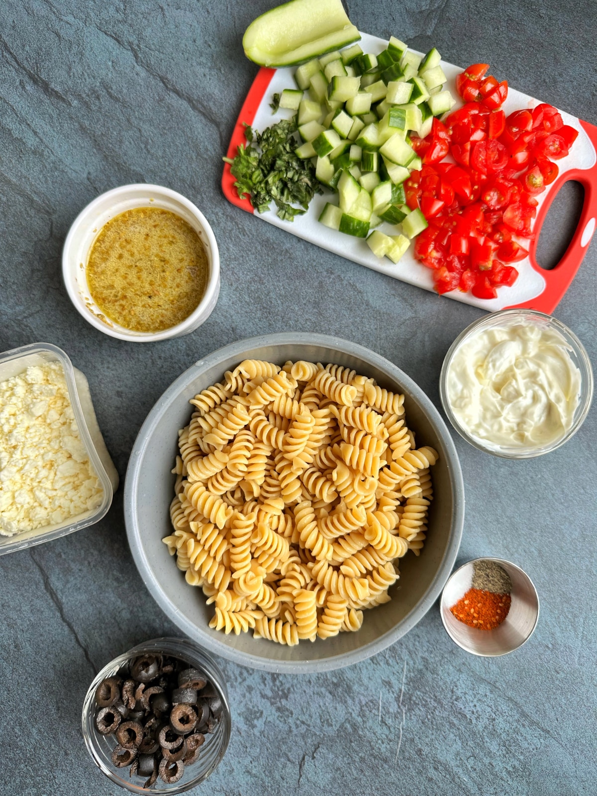 A bunch of bowls and cutting boards filled with Greek pasta salad ingredients, before combining together.