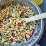A large bowl of Mediterranean Pasta Salad mixed together with a wooden spoon in the bowl.