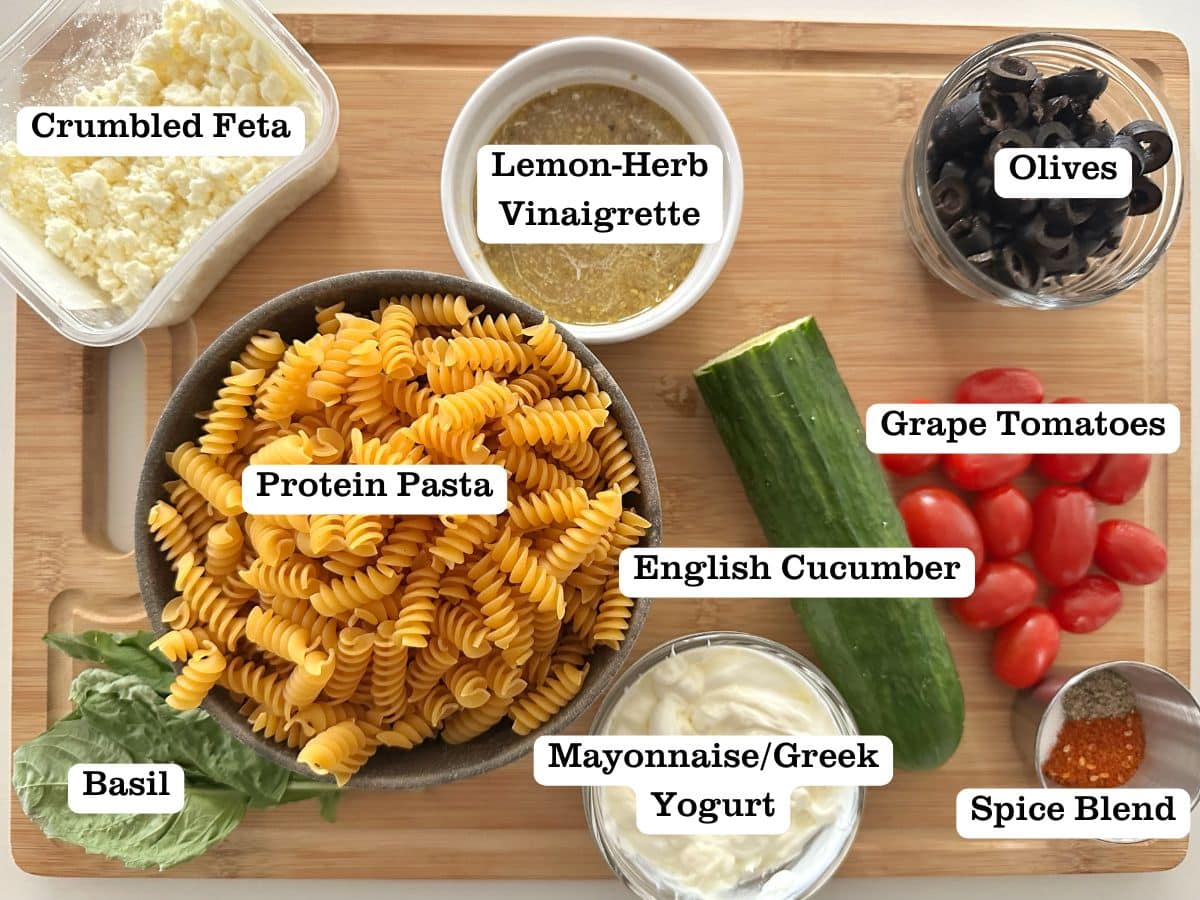 A wooden board containing bowls of ingredients for Greek pasta salad.