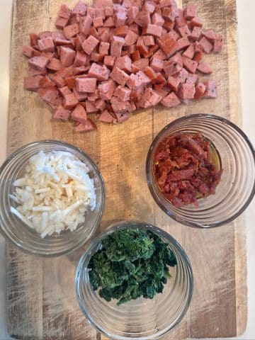 A wooden board containing diced sausage and bowls of frozen potatoes, spinach, and sun-dried tomatoes.