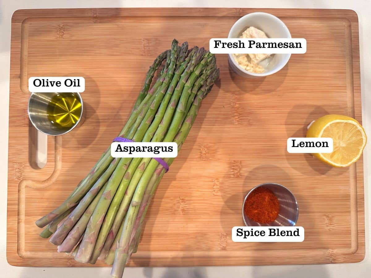 A wooden board containing ingredients for roasted asparagus, including parmesan and a fresh lemon.