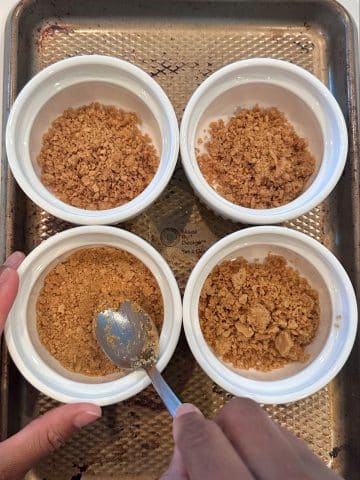 A hand using a spoon to push graham cracker crusts into small ramekins.