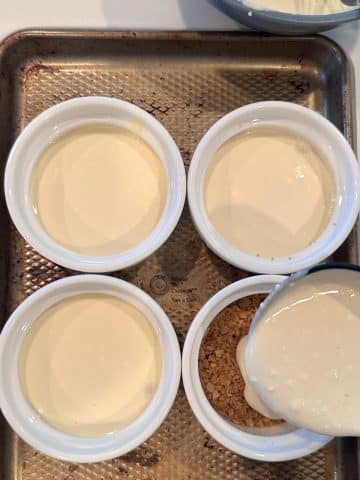 An off-screen hand ladling cheesecake filling onto a ramekin with a graham crust inside.
