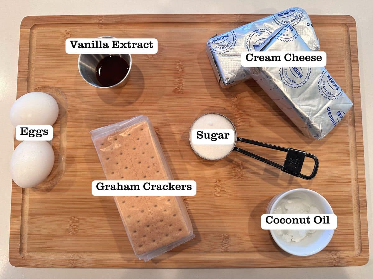 A wooden board containing bowls of ingredients for mini cheesecakes, including eggs, graham crackers, and coconut oil.