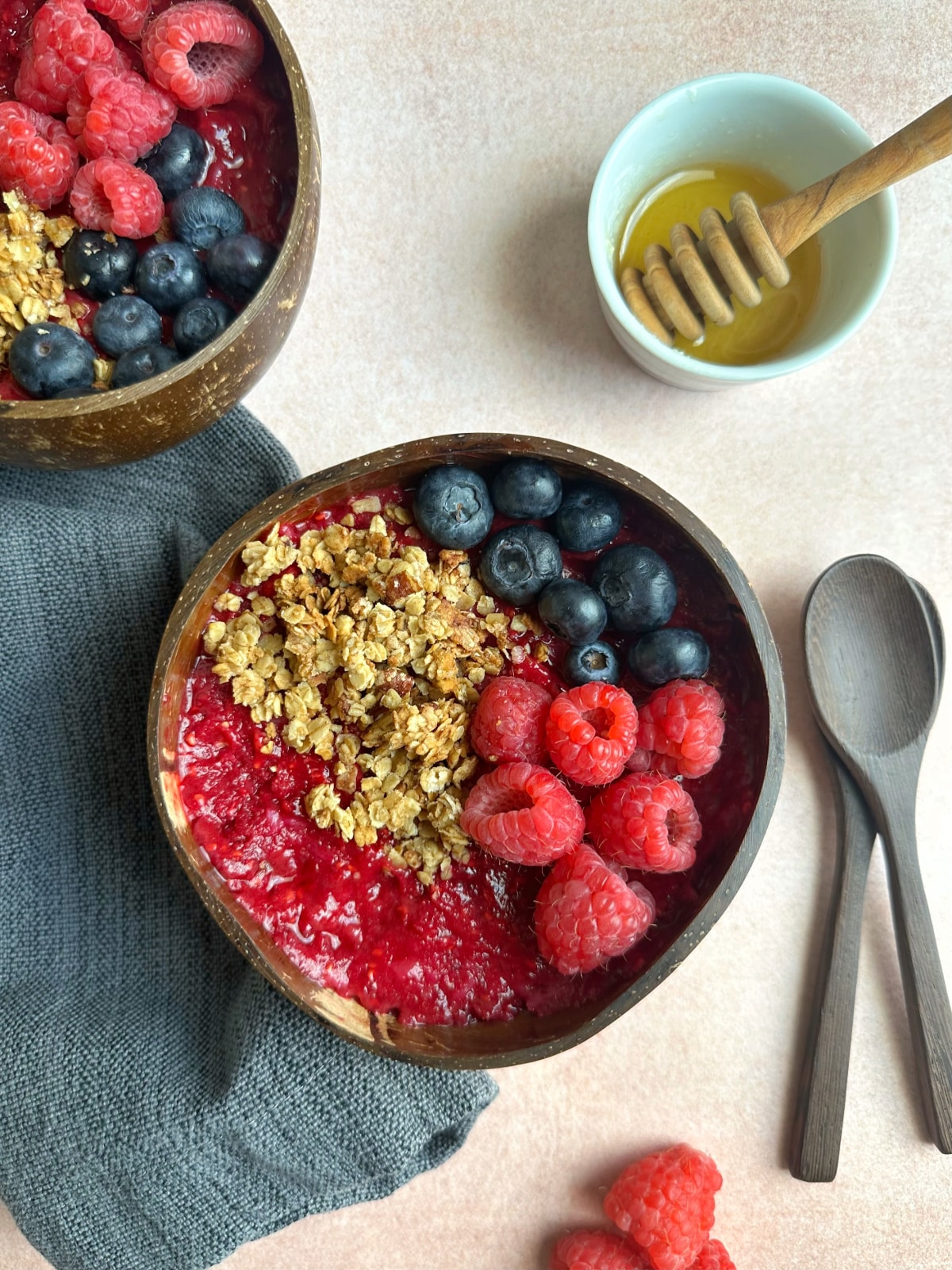 Two bowls of raspberry smoothie topped with granola, fresh blueberries, and fresh raspberries.