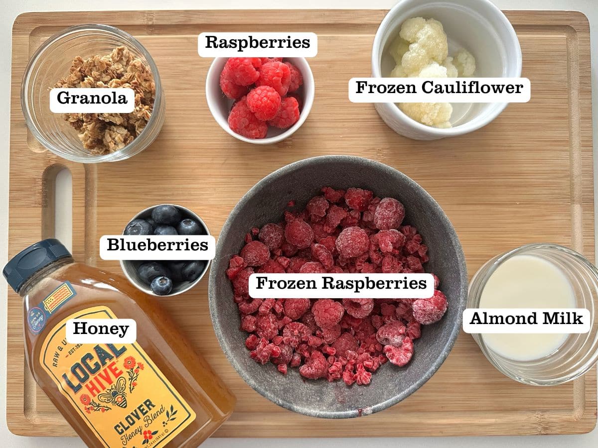 A wooden board containing ingredients for raspberry smoothie bowls, including frozen fruit, almond milk, and honey.