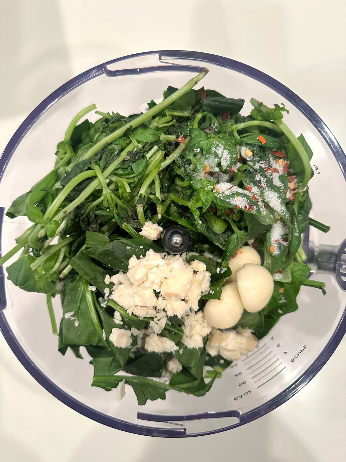 A food processor containing fresh spinach leaves, basil leaves, garlic, and parmesan cheese.