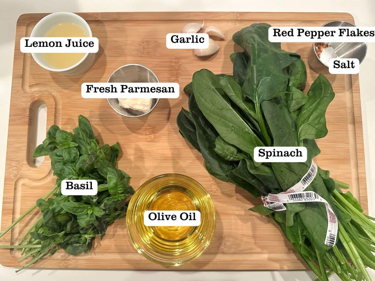 A wooden board containing the ingredients for nut-free pesto, including spinach leaves, bowls of olive oil, lemon juice, and fresh parmesan cheese.