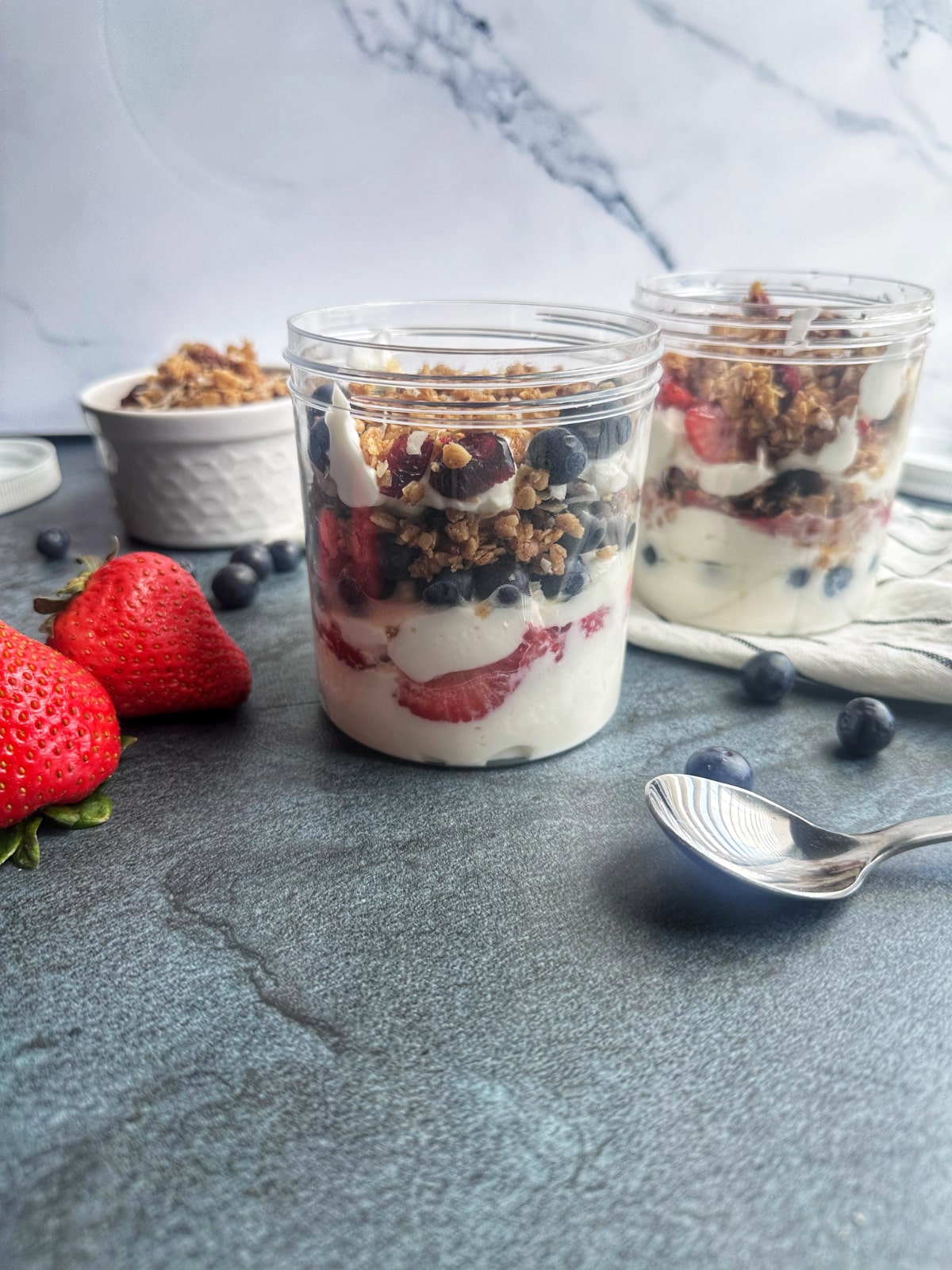 Two plastic jars filled with layered greek yogurt, berries, and honey, with extra ingredients displayed on the side.