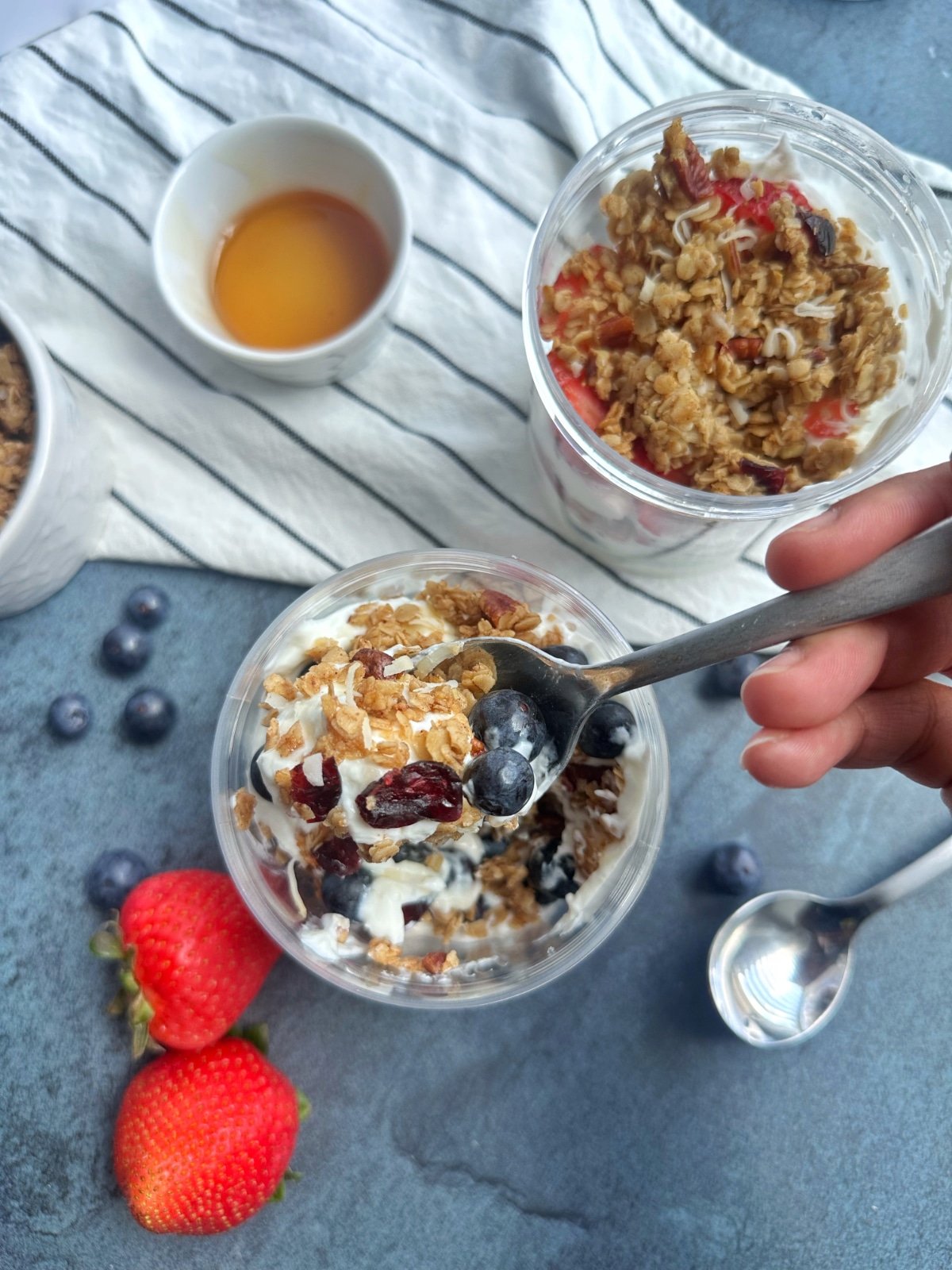 A hand scooping a spoonful out of a container filled with greek yogurt, blueberries, and granola.