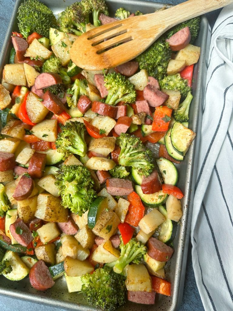 A big pan of Sheet Pan Sausage and Veggies with a white and black striped towel to the side.