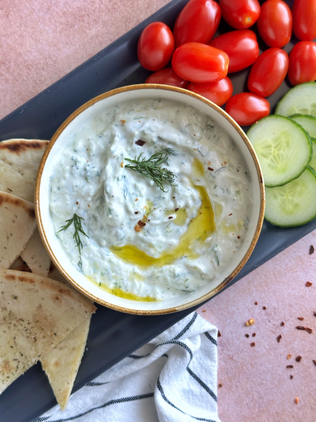 A bowl of tzatziki sauce with an olive oil drizzle on a platter with dippable veggies and bread on the side.