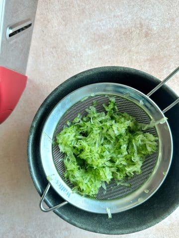 Grated cucumber in a fine mesh sieve over a bowl, to drain the liquid.