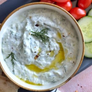 A bowl of homemade tzatziki surrounded by dipping foods like grape tomatoes and sliced cucumber.