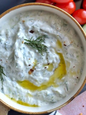 A bowl of homemade tzatziki surrounded by dipping foods like grape tomatoes and sliced cucumber.