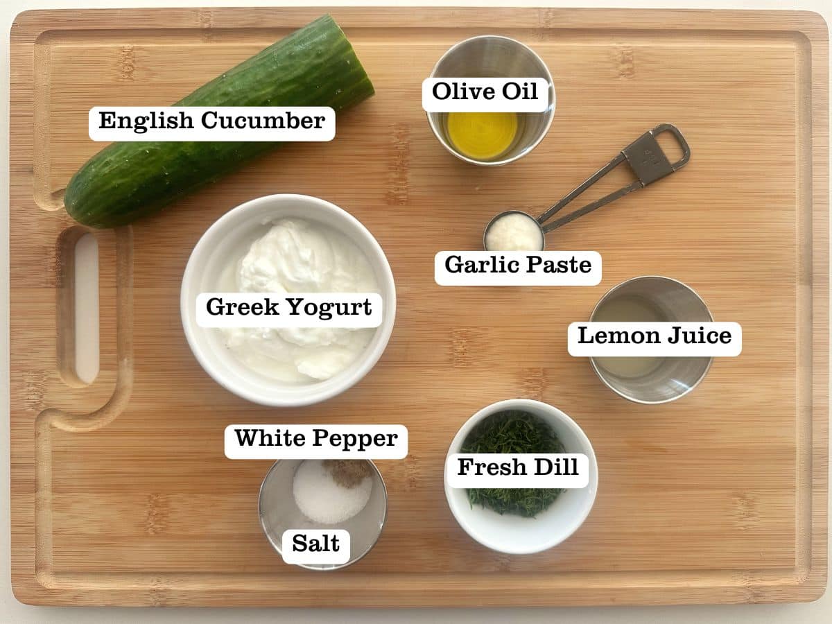 A wooden board containing bowls of ingredients for tzatziki, including Greek yogurt and cucumber.