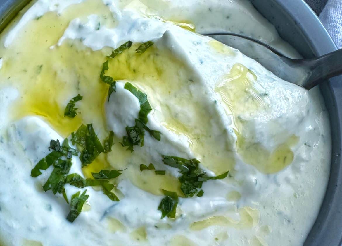 A spoon scooping up homemade Greek tzatziki sauce out of a grey bowl.
