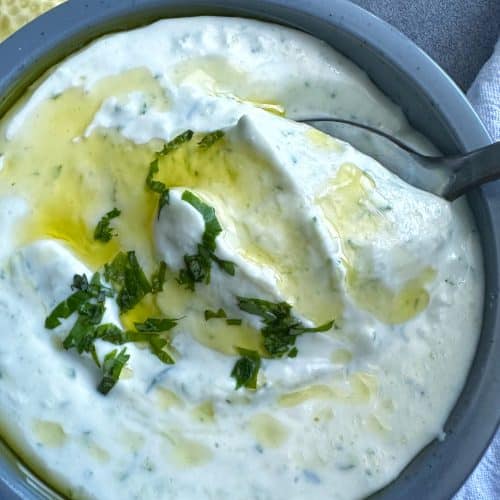 A spoon scooping up homemade Greek tzatziki sauce out of a grey bowl.