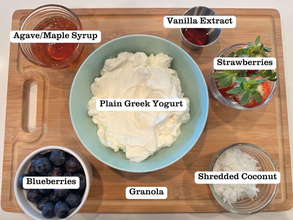 A wooden board containing bowls of yogurt, berries, coconut shreds, syrup, and vanilla extract.