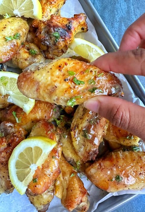 A hand holding a lemon pepper wet wing with more on a tray in the background.