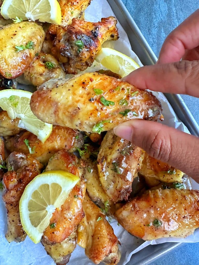 A hand holding a lemon pepper wet wing with more on a tray in the background.