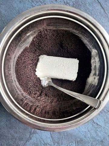 A block of cream cheese in the middle of a bowl of crushed chocolate sandwich cookies.
