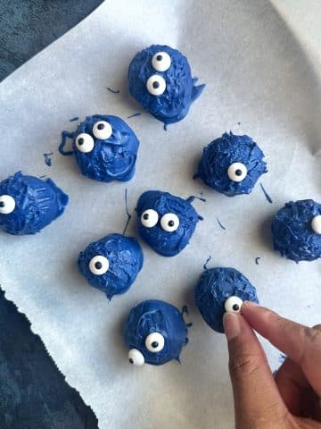 A hand placing mini candy eyeballs onto blue, chocolate covered truffle balls.