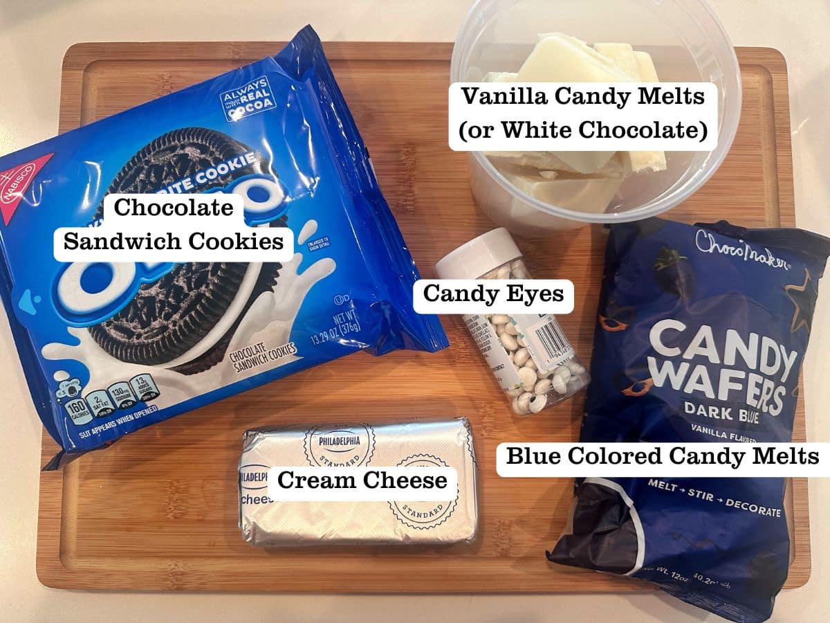 A wooden board containing a package of Oreo cookies, blue candy wafers, a deli container filled with vanilla candy melts, cream cheese, and mini candy eyeballs.