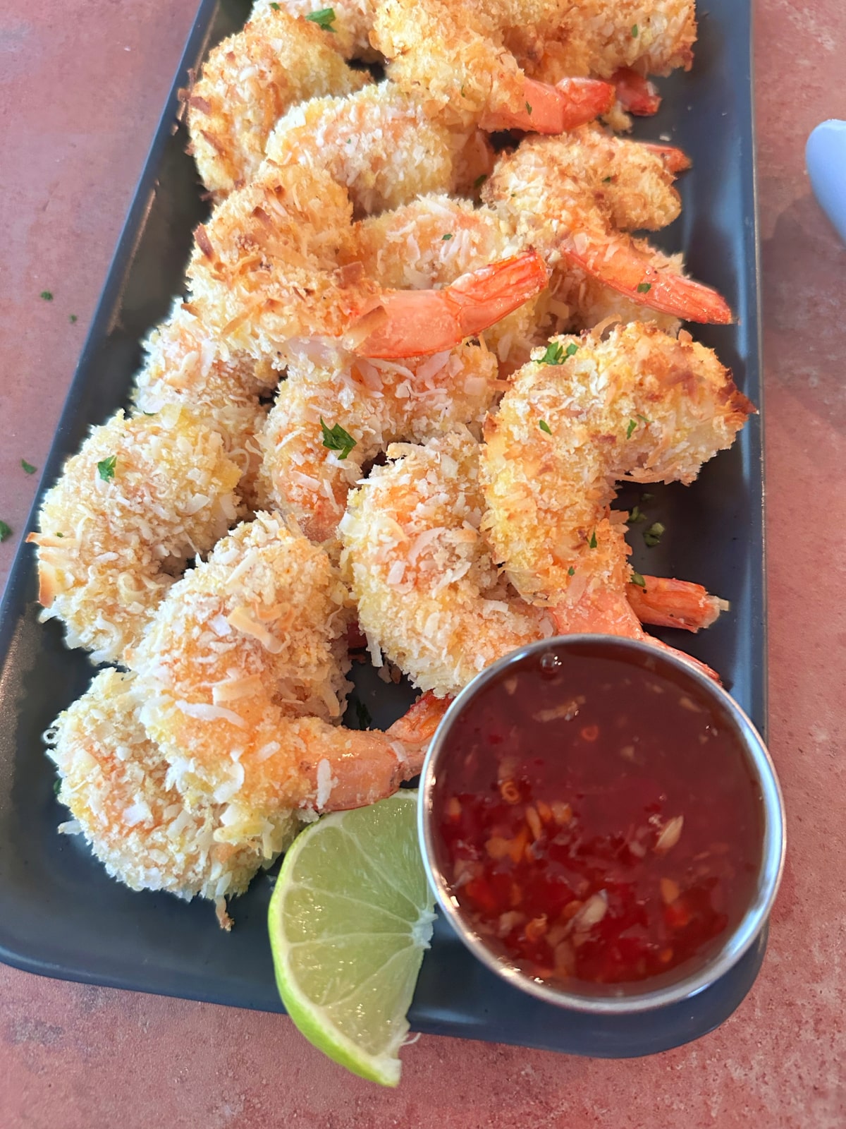 A pile of coconut shrimp on a serving tray with sweet chili sauce and lime on the side.