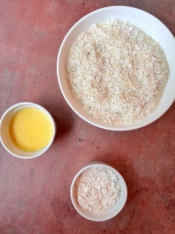 Three separate bowls at a dredging station, with whisked eggs, flour, and shredded coconut in each bowl.