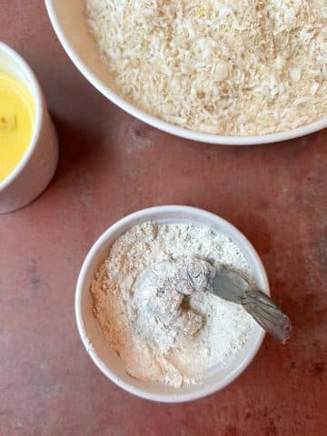 A jumbo shrimp with its tail on, dipped and covered in a bowl of flour.