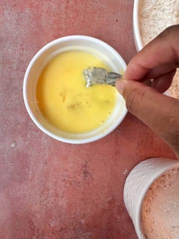 A hand dipping a singular shrimp into a bowl of whisked eggs.