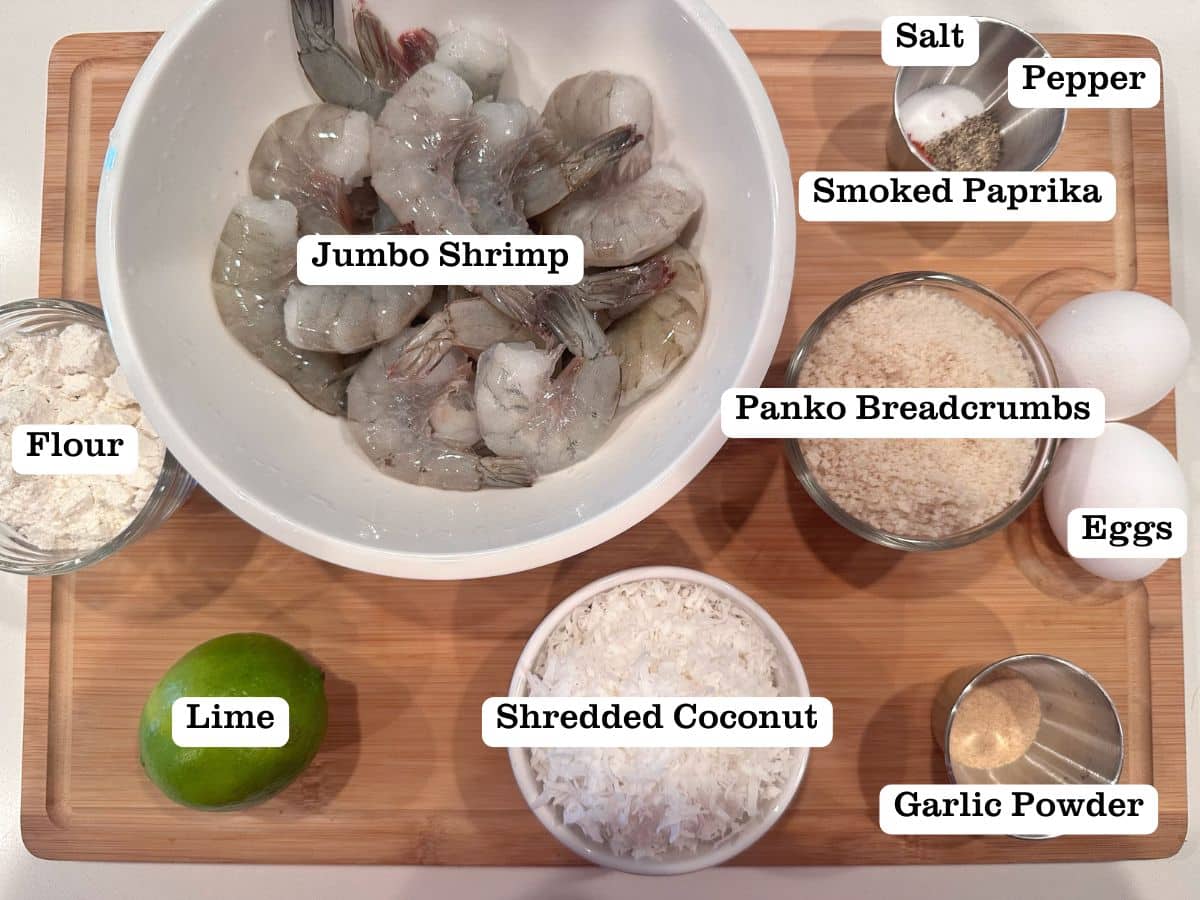 A wooden board containing ingredients for Coconut Shrimp, including panko breadcrumbs, eggs, and lime.